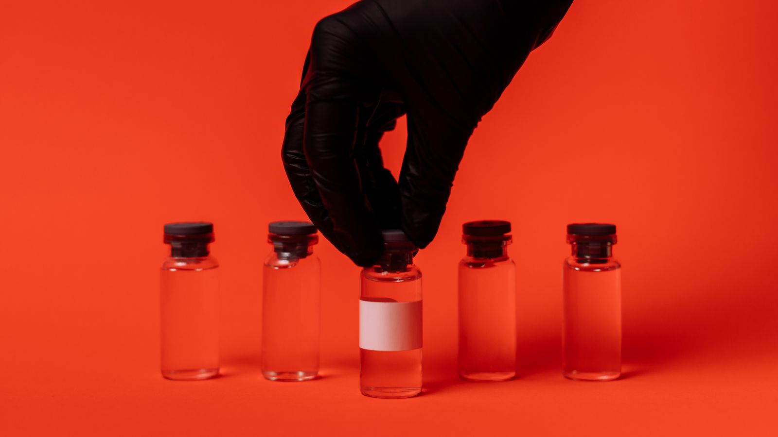 A gloved hand holding a small glass vial against a bright red background.
